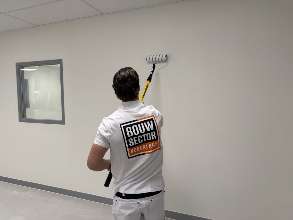 Schilder in een Bouwsector Nederland-shirt rolt een witte wand onder een plafond van platen