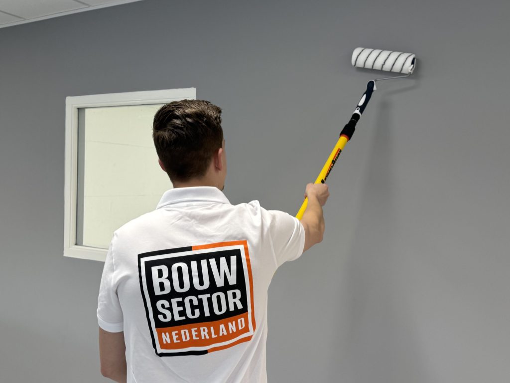 Schilder werkt een grijze muur bij, met links een raam in een wit gelakt houten kozijn