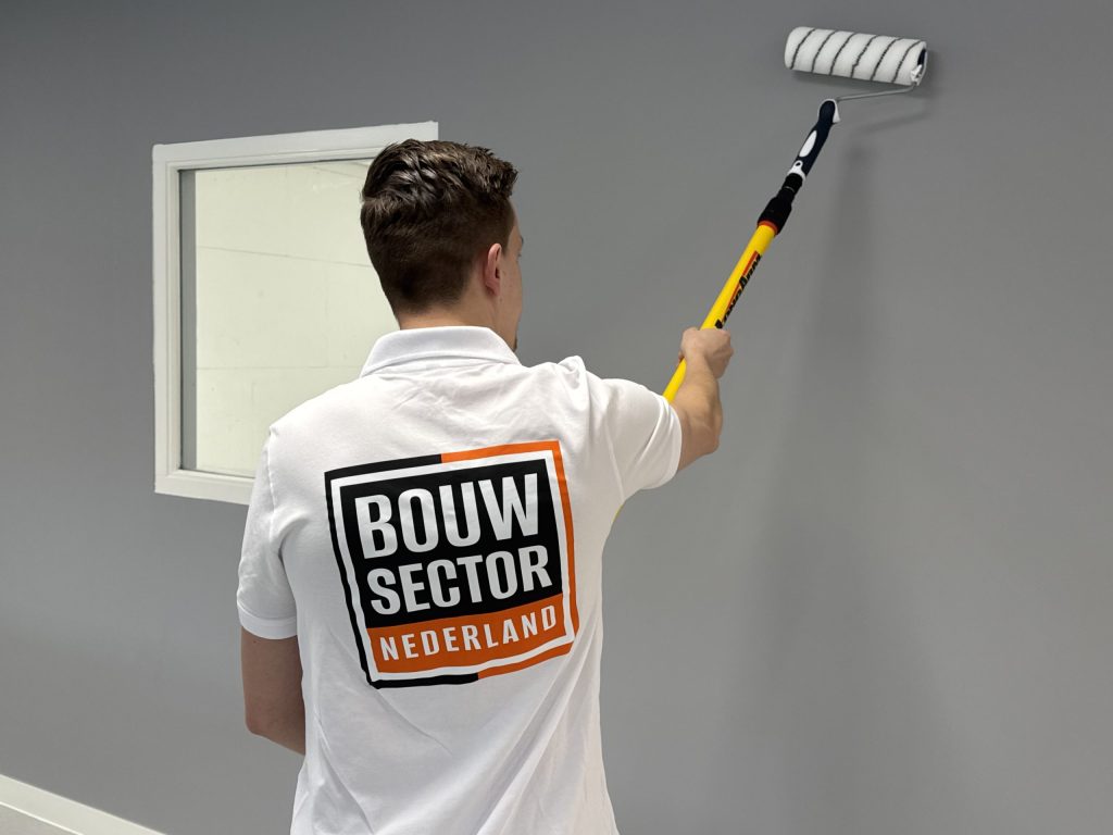 Schilder met het logo van Bouwsector Nederland op zijn polo rolt een grijze wand naast een wit gelakt raamkozijn