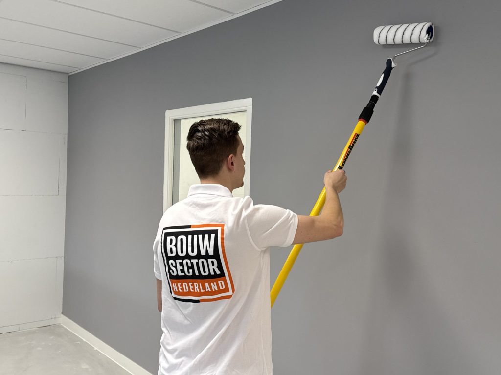 Schilder rolt met een gele telescoopsteel een grijze wand tot onder het plafond, naast een deur met wit kozijn