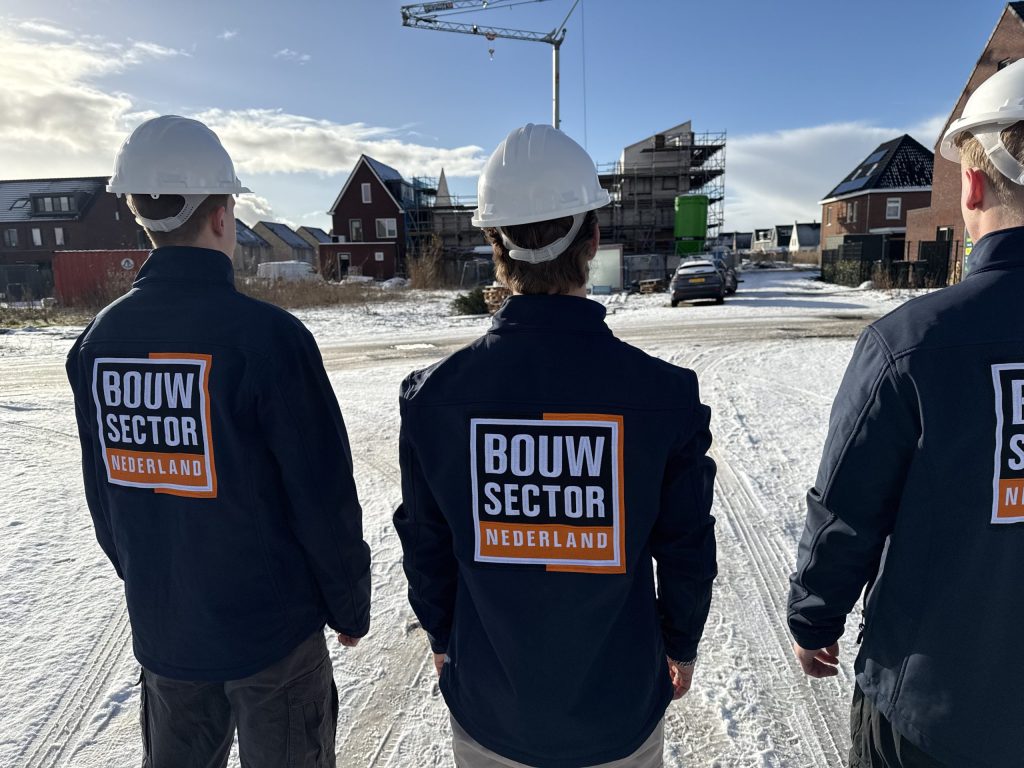 Drie vakmensen van Bouwsector Nederland in blauwe jassen naast elkaar op een bouwplaats met hijskraan