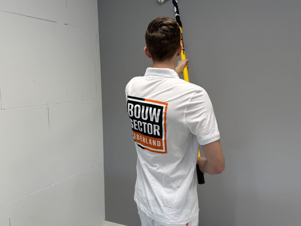 Schilder in wit poloshirt rolt een grijze muur naast een kale wand van kalkzandsteen